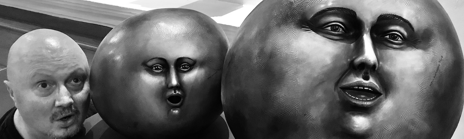 Me (with my bald head) in a black t-shirt bending down beside an end table with two different sized round balls with faces on them (modern art) in the main lobby at the Excellence Riviera Maya, Cancun, Mexico.