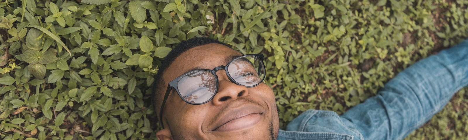 A man lies on the plant covered ground with his eyes shut and a happy grin on his face. He looks relaxed, he’s not worried about the tax deadline