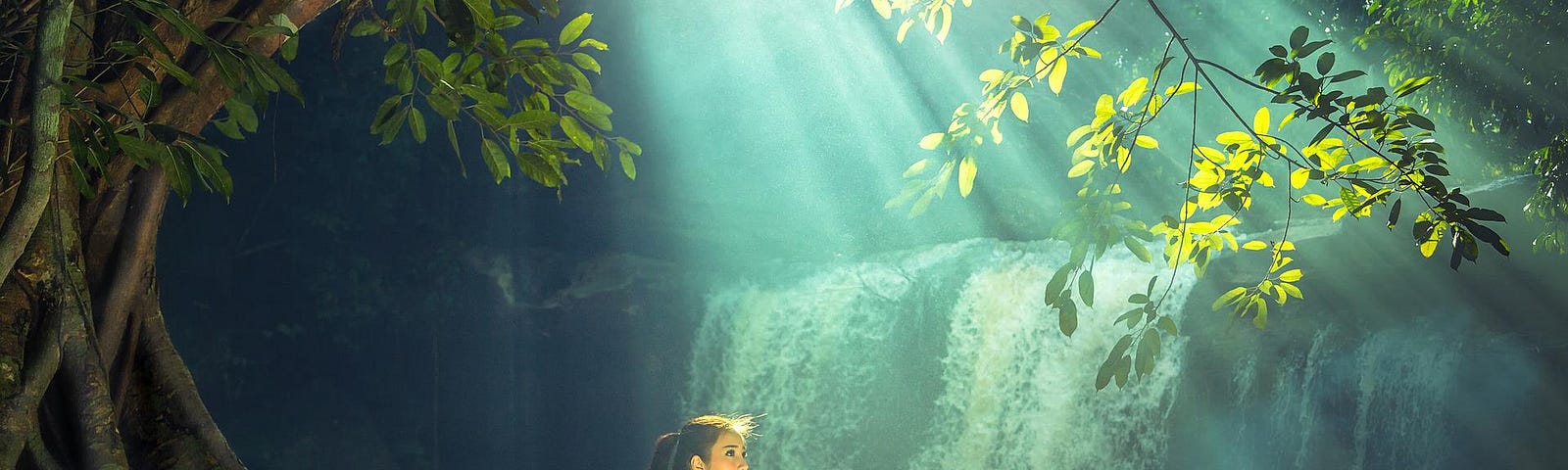 In a forest, a young woman sits on a root of a large tree that bends over her head beside a waterfall. Sun rays shine through the leaves above her head.