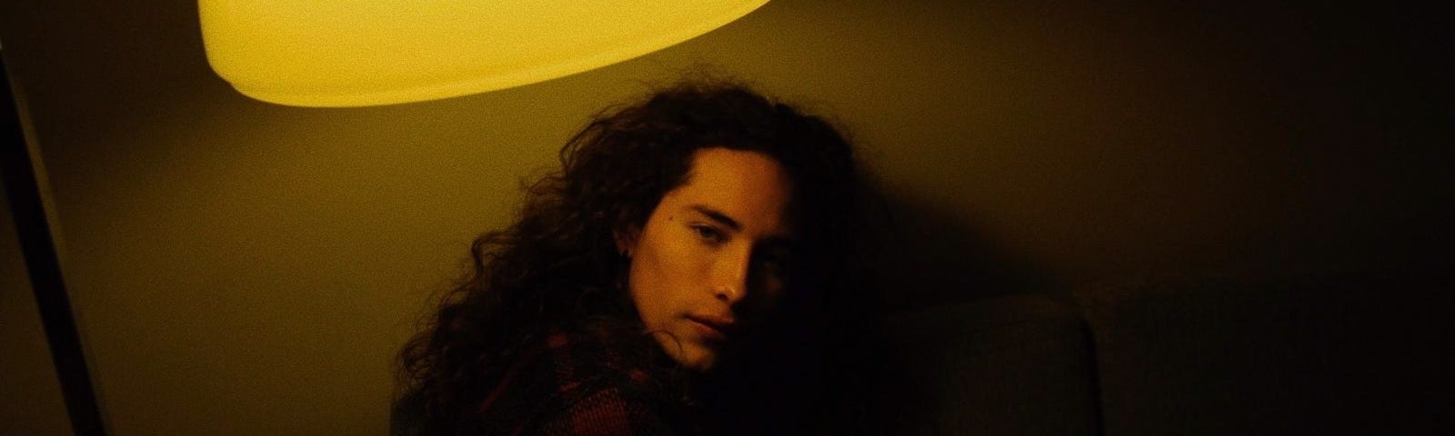 A man with long dark hair stares at the camera beneath a dim light.