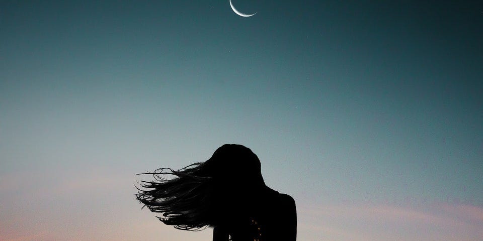 The sihlouhette of a woman standing under a crescent moon looking like she’s in a trance.