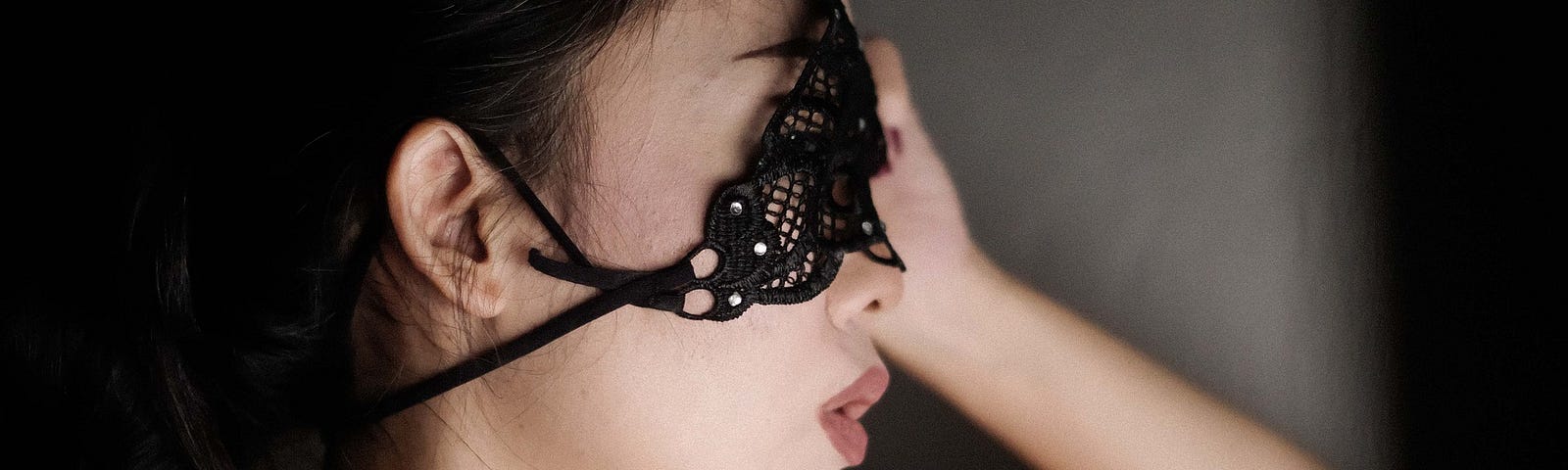 Image of woman, brunette hair, red shoulder straps, wears black masquerade mask, pouting lips