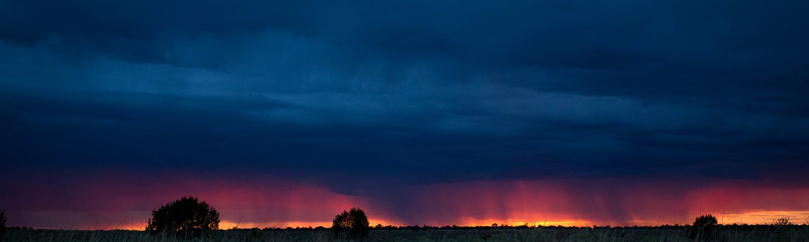 Dark, heavy clouds above an orange ribbon of sunset with a dark field in the foreground. (opacarophile, and pluviophile)