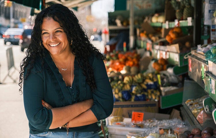 Ivelyse Andino, an Afro-Latina woman with long dark curly hair, stands in front of an open air market, with her arms crossed, smiling, wearing a blue three-quarter sleeve shirt, two silver necklaces, and blue jeans.