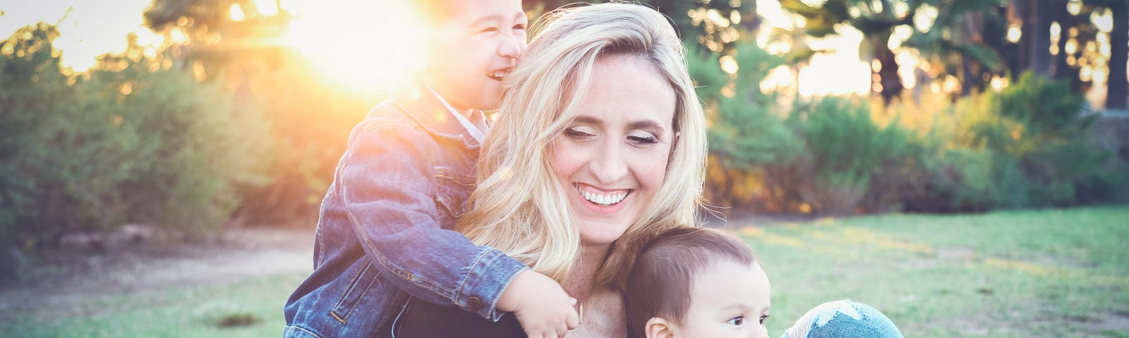 A woman sitting on the grass holding a toddler in front of her and smiling. A kid standing behind her smiling and holding her shoulder.