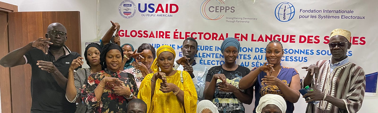 A group of people in front of a USAID banner pose for a picture while using sign language to display the word “vote” with their hands.