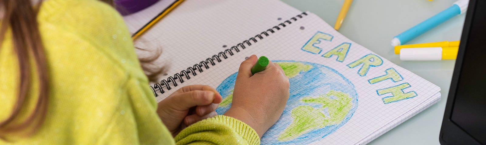 A child wearing a yellow sweater is drawing the planet Earth on a notebook. The word “Earth” is colored in green.