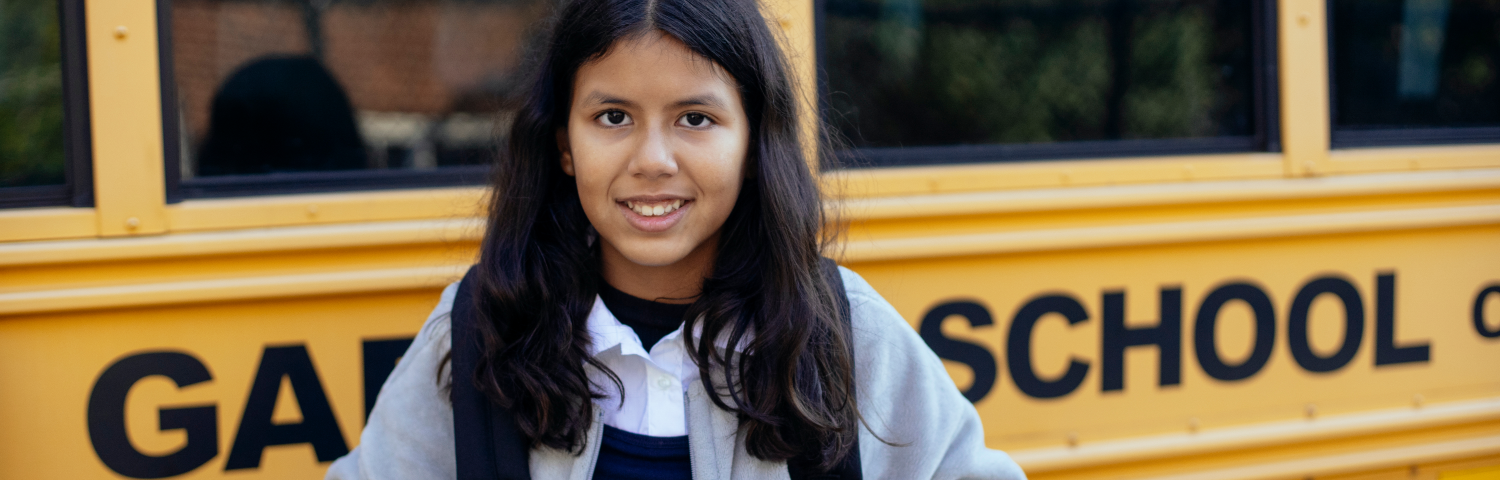 A preteen girl stands smiling in front of a school bus.