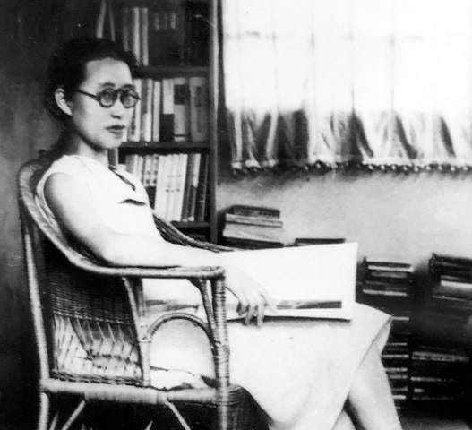 Sagawa sitting in a chair in a white dress with a book on her lap, her hair in a bun, wearing glasses.