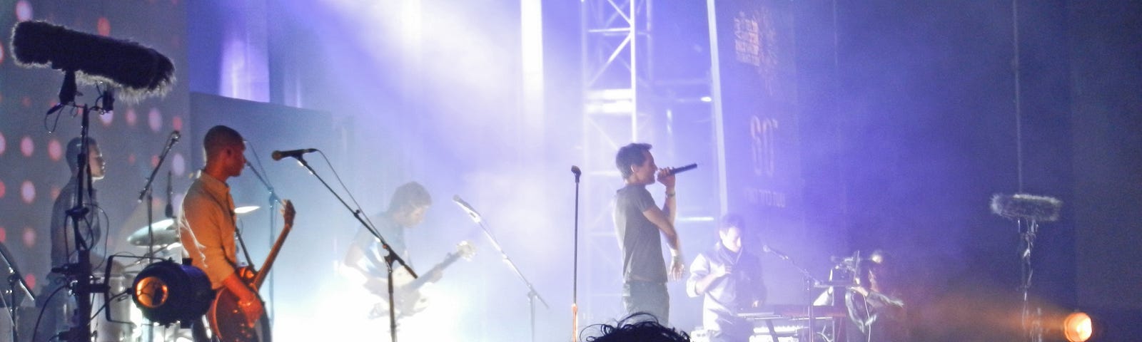 Concert stage — Base player on the left side of the stage and the singer is in the middle holding a microphone up to his mouth. Fans are in the foreground and bright white lights are behind the band.