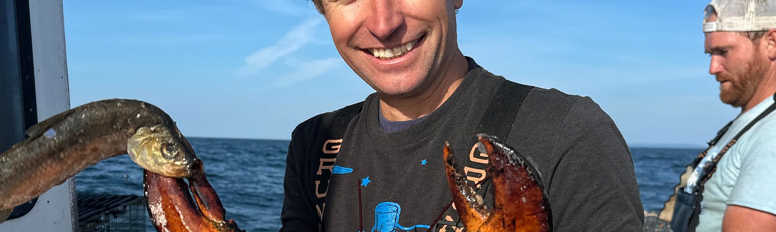 Author is holding a giant lobster. he is wearing soiled working overalls and gloves. A pile of pigskins is to the side — it will be used for bait.