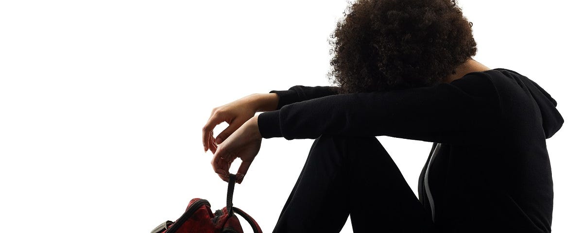 Young person of color sits with a backpack in a posture of sadness, with head between arms.