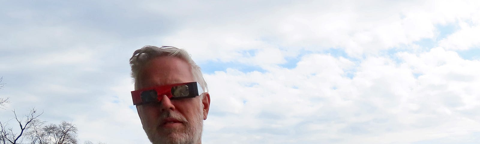 Man wearing red glasses with very dark lenses. Clouds are overhead.