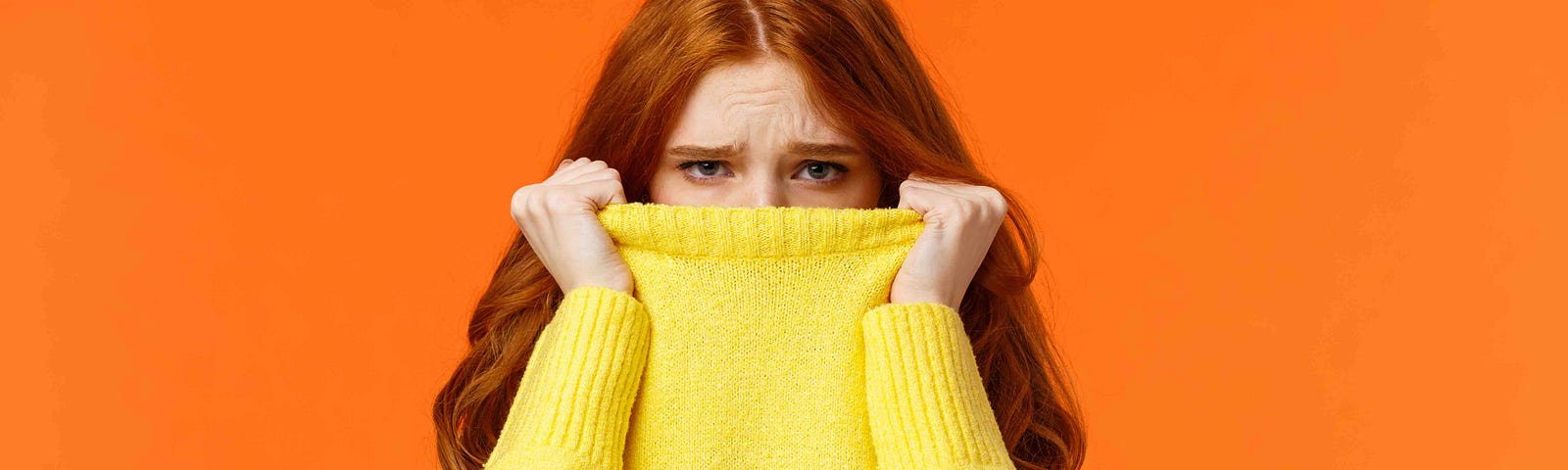 A photo of an embarrassed woman hiding her face in a yellow sweater against an orange background.