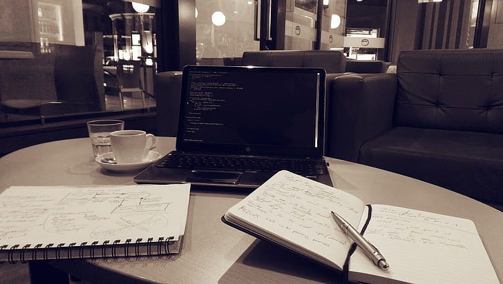 A laptop, showing some code, next to a cup of coffee and two notebooks