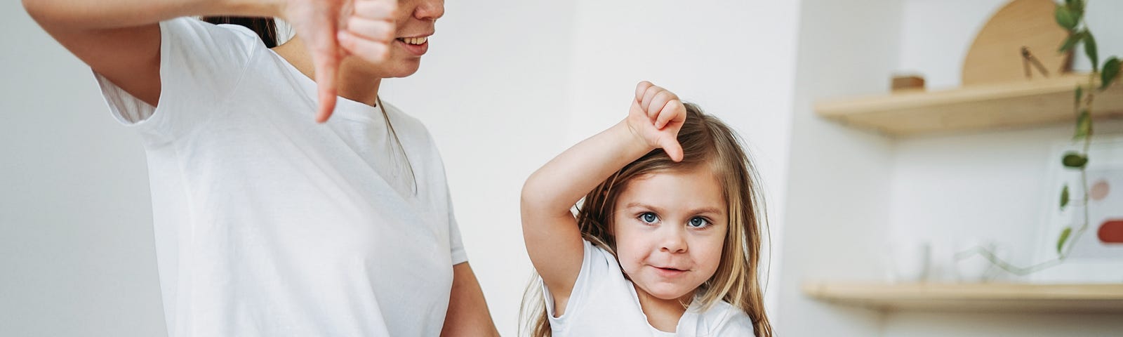 A child gives a thumbs down to her mother who sits beside her on a white bed.