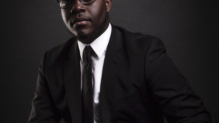 Professional Black businessman in dark suit and glasses sitting confidently against black background — corporate headshot photography