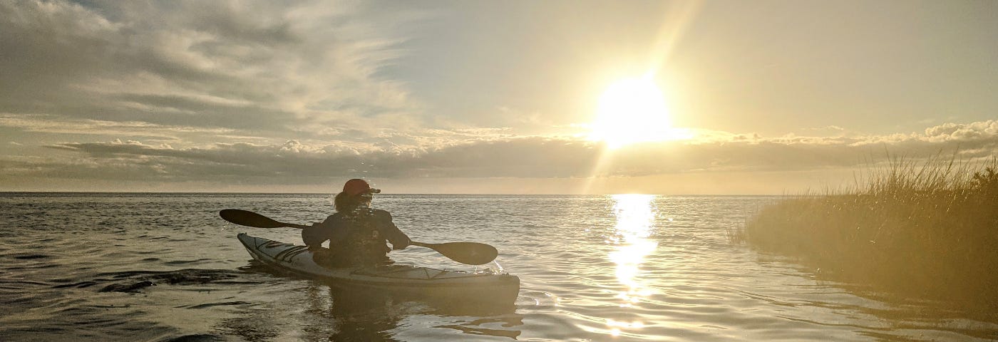 A woman paddles a kayak on a calm, sunlit ocean.