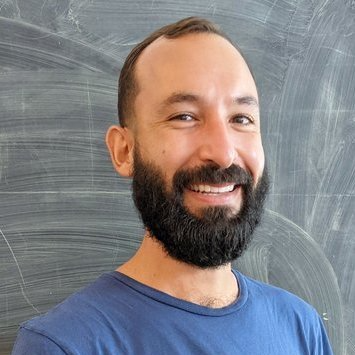 A man with short brown hair and a black beard, wearing a dark blue shirt, smiles while standing in front of a dirty chalkboard.