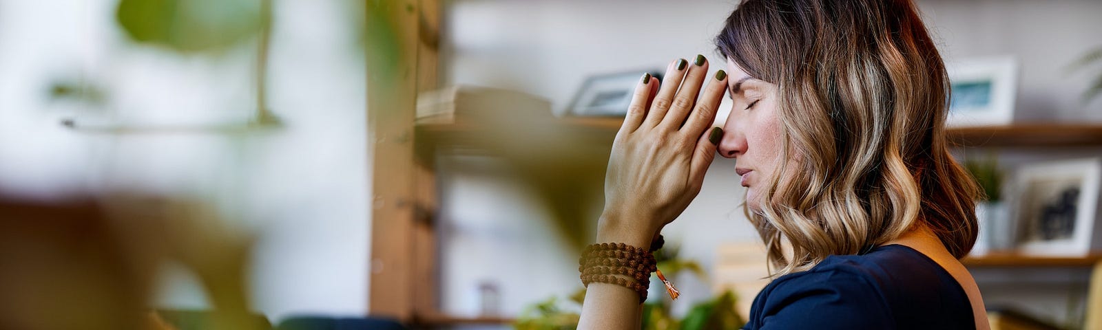 A woman in a dark blue shirt in her room with her hands in a prayer shape and mala beads around her wrist