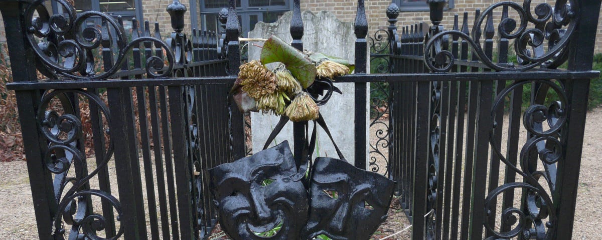 The grave of Joseph Grimaldi. A weathered headstone and grave is surrounded by black, ornate, cast-iron railings. The twin masks of tragedy and comedy — also in black iron — hang on the railing.