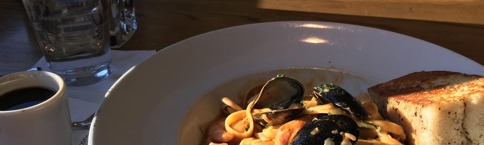 Seafood pasta with a piece of bread on the side. And a coffee next to the dish.