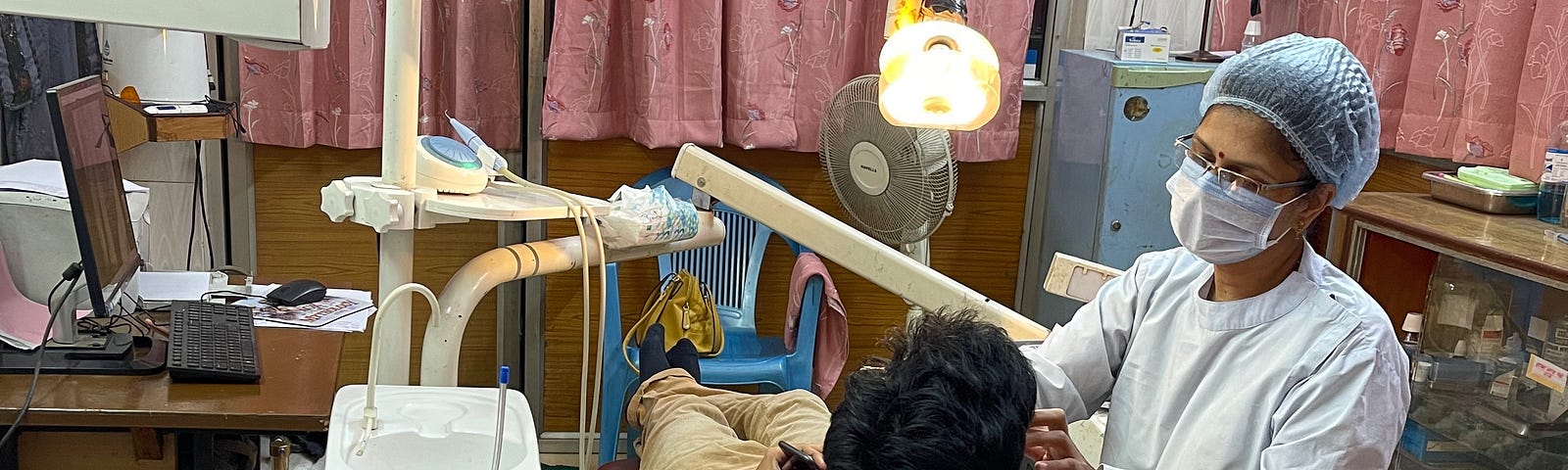 A dentist at work on a patient. She is dressed in a dentist’s apron. She is wearing a face mask and a disposable scrub cap. She has on harem pants and a long shirt under her apron.