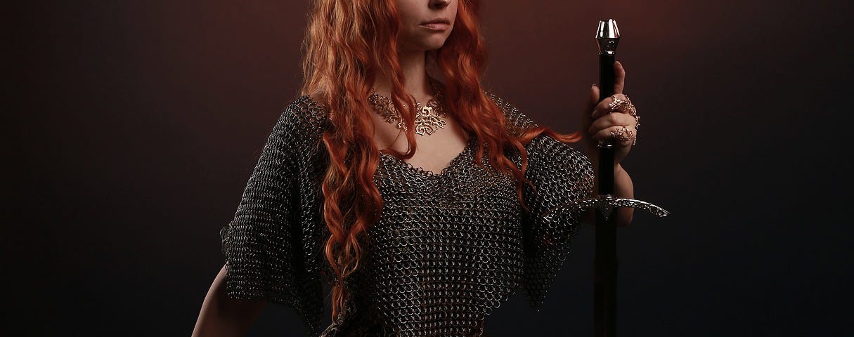 A woman with long red hair wearing a crown and a long dark dress is on one knee holding a sword.