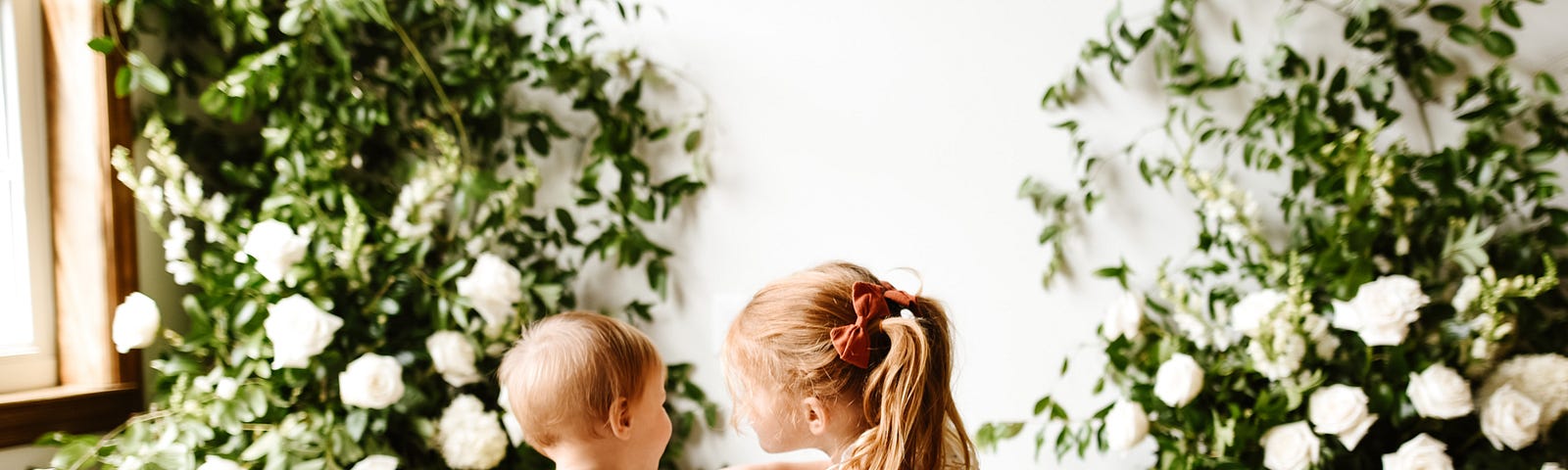 Little girl holds hands of baby while both sit on the floor with white roses in the background