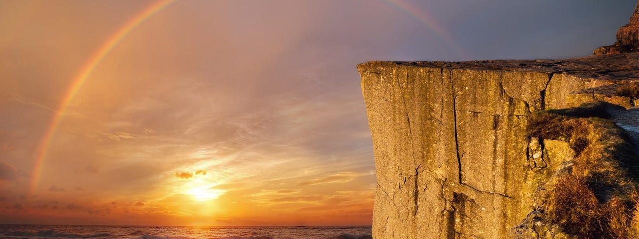 A rainbow arcs over the sun rising over the ocean. In the foreground, a sharp yellow cliff rises above the rocky beach.