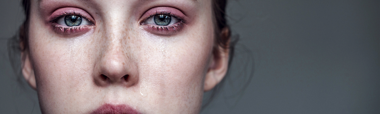 A close up of a young woman with light skin, freckles and green eyes. Her eyes are red and swollen and tears stream down her face.