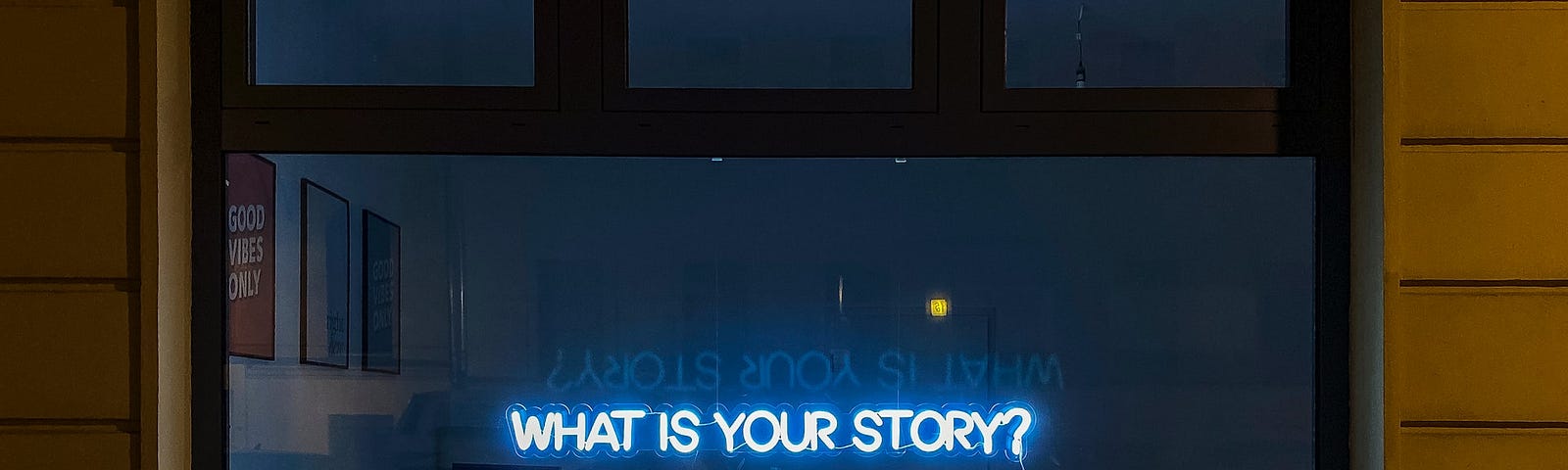 An exterior view of a structure with a large window peeking into an unlit modern office. A neon sign in the window reads: WHAT IS YOUR STORY?