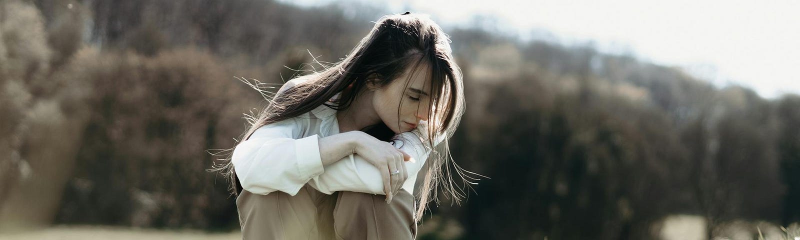 A woman with long brown hair sits in a field of dry grasses, arms folded on her knees and glancing downwards.