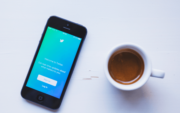 A phone with the twitter app on the screen and a cup of coffee.
