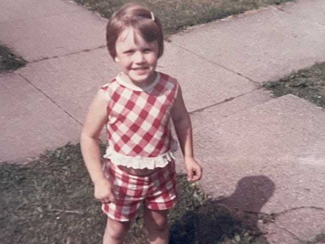 Little girl in red & white checked short and top, smiles up at the camera