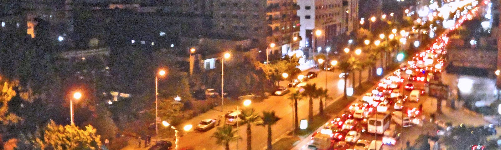 Nighttime heavy traffic on a wide 8-lane two-way road. Buildings are on both sides of the street. There are palm trees on the boulevard between traffic.
