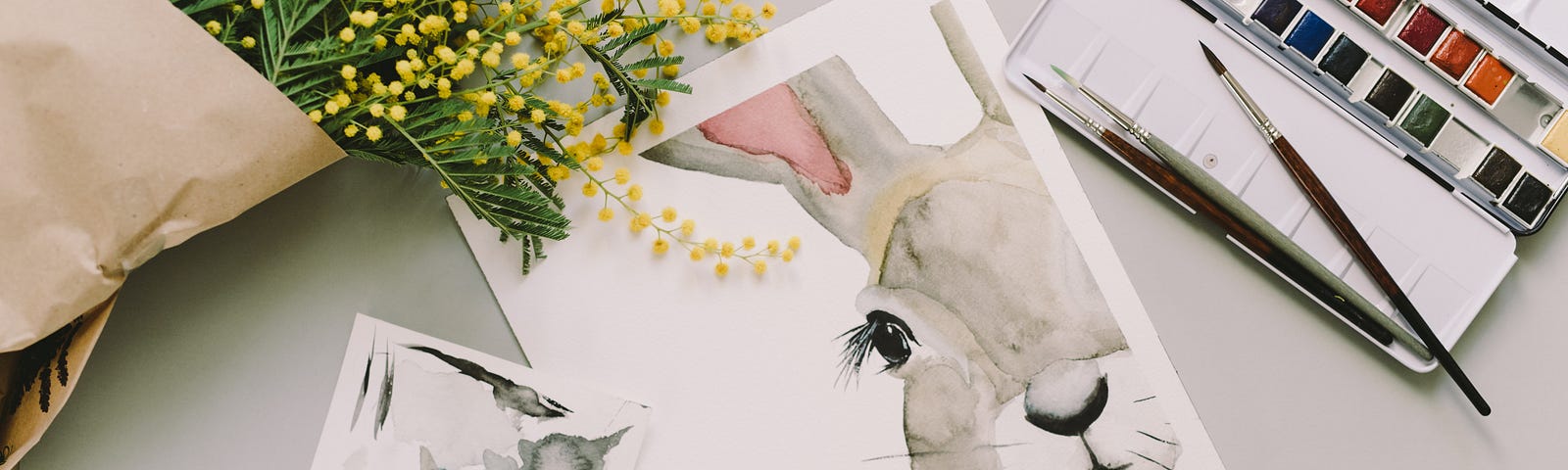 A watercolor painting of a bunny sits on a white table next to yellow flowers.