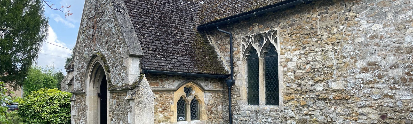 A Brompton bicycle sitting on a path outside an old stone church. The sky is blue. The grass is green. There is a little raised flower bed in the foreground. The flowers have yet to bloom to their full potential.