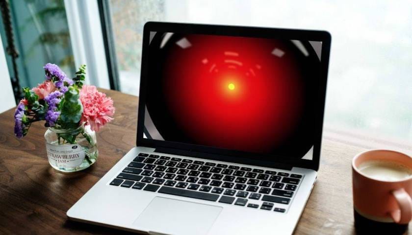 A laptop on a desk; the laptop’s screen is filled with the glowing red eye of HAL 9000 from 2001. Image: Cryteria (modified) https://commons.wikimedia.org/wiki/File:HAL9000.svg CC BY: https://creativecommons.org/licenses/by/3.0/deed.en