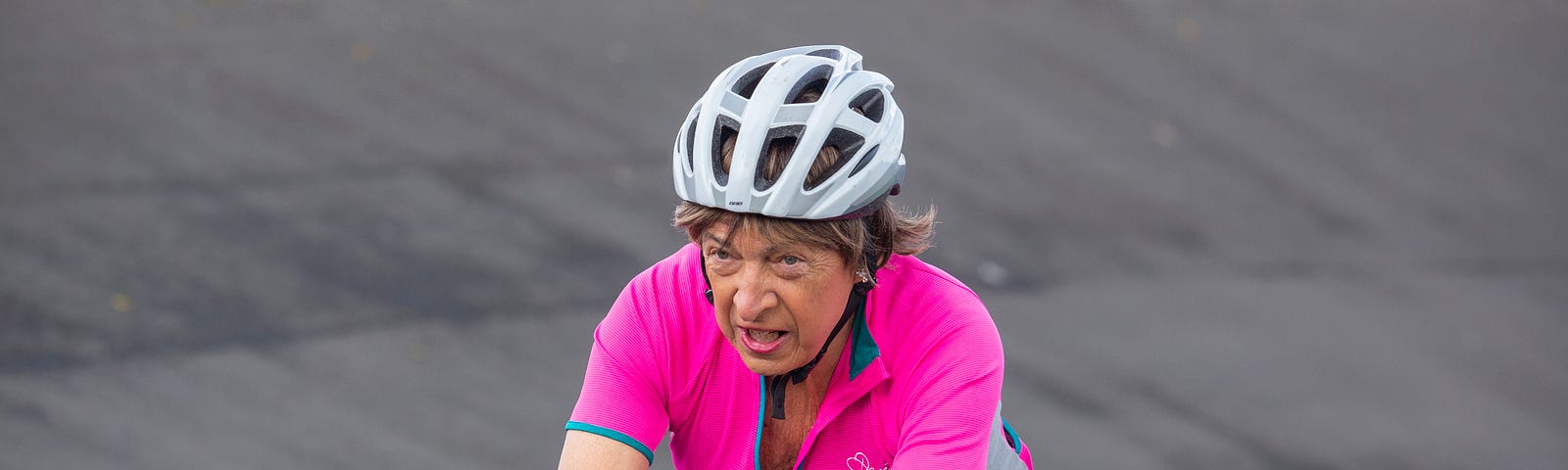 Michaela is cycling. She wears a pink cycling top, black shorts and a helmet as she rides round a track.