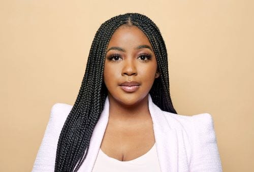 Ashlee is a Black woman centered in the frame from the chest up. She has long braids, half pulled over her right shoulder and trailing off camera, the other half behind her left shoulder. She is wearing a white blouse and blazer and sits in front of a light pink background.