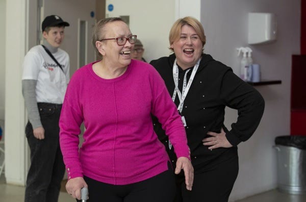 An older woman grins to someone off camera as she dances. A younger woman stands behind her laughing with her hands on her hips as she watches on.