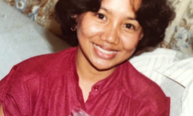 A smiling brown-skinned woman wearing a burgundy blouse while holding and feeding a baby.