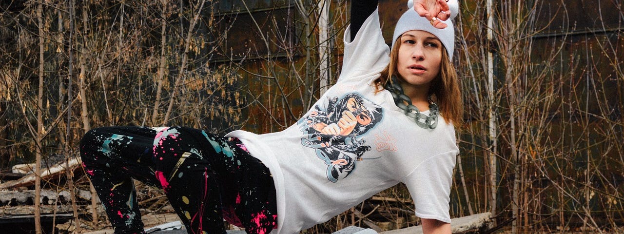A young girl in hip clothes — beanie, tshirt, leggings, boots and glove — in a breakdancing pose.