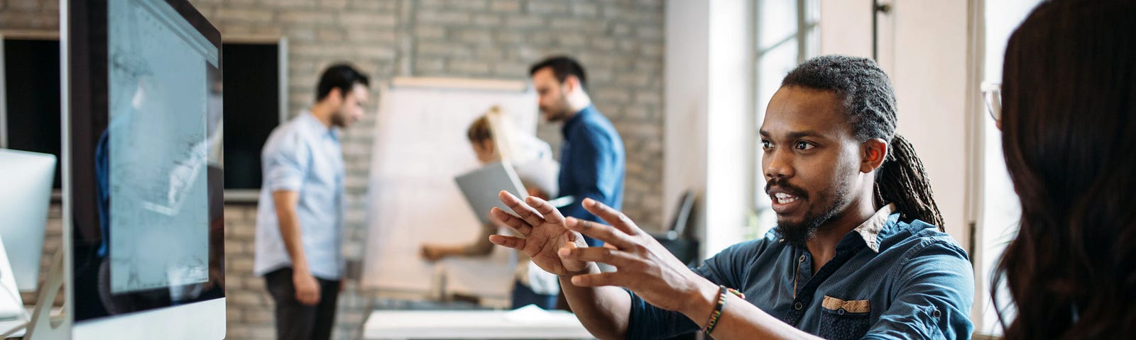 Two people sit at a desk, looking at a computer screen. One person points, while the other has their hands in their lap.