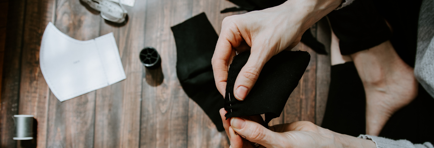 hands holding a piece of black fabric with needle in the fabric, crossed legs in background on wood floor with more fabric pieces, thread, and measuring tape