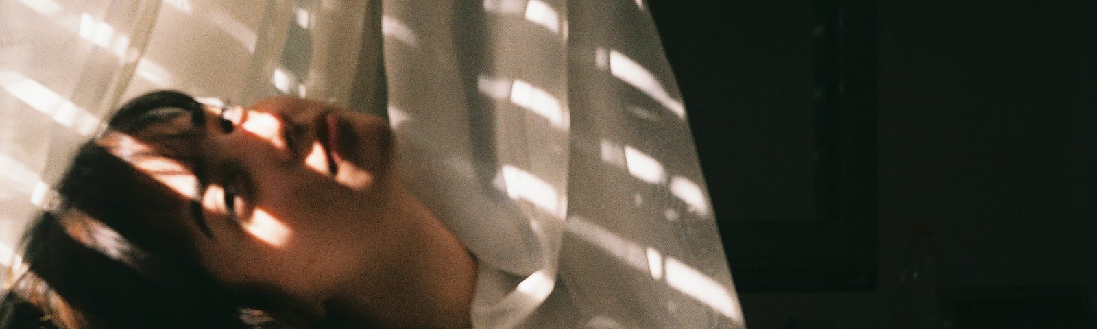 A young woman resting her head on a windowsill, a white curtain half draped around her neck and back of her head. Light shining in on her face.