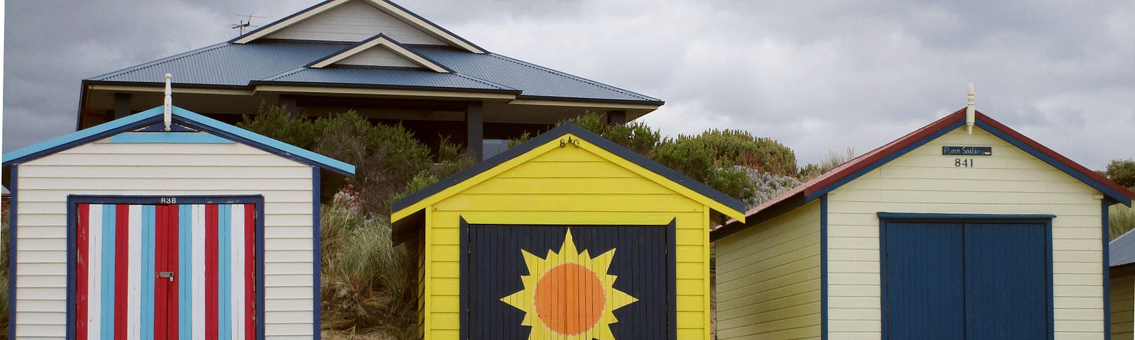 Three colorful bathing boxes on the beach with the roof line of a house and grey clouds in the background.