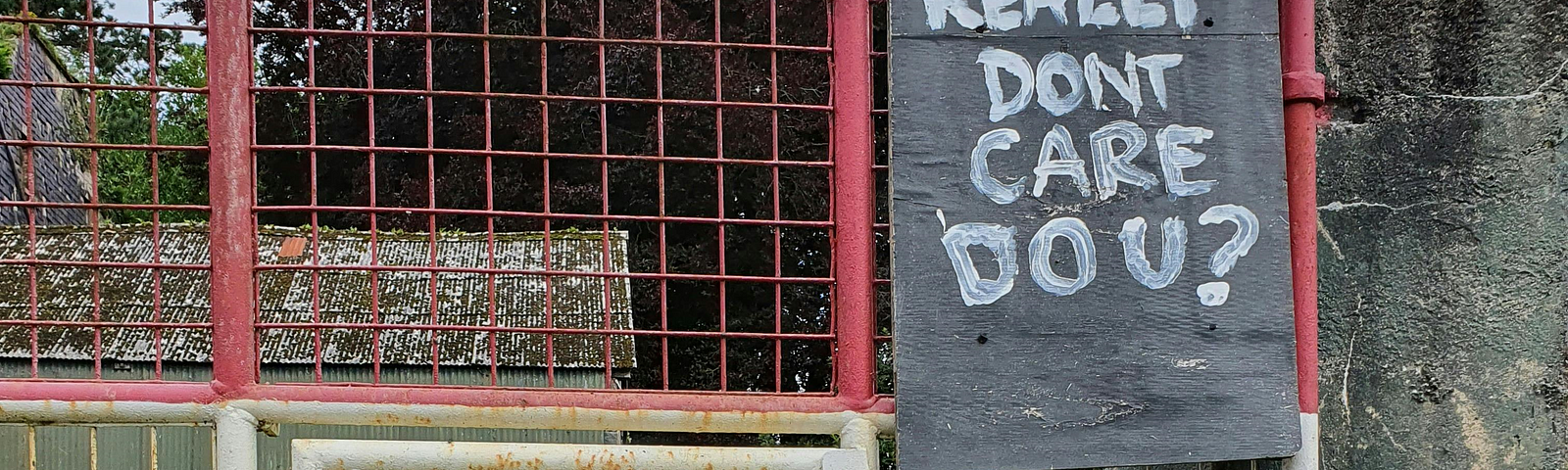 A metal fence that you can see through and looks like a farm building behind it. There is a cut out painting that looks like Donald Trump to one side with a big chalk board above his head. On the board is written, ‘I really don’t care. Do you?’