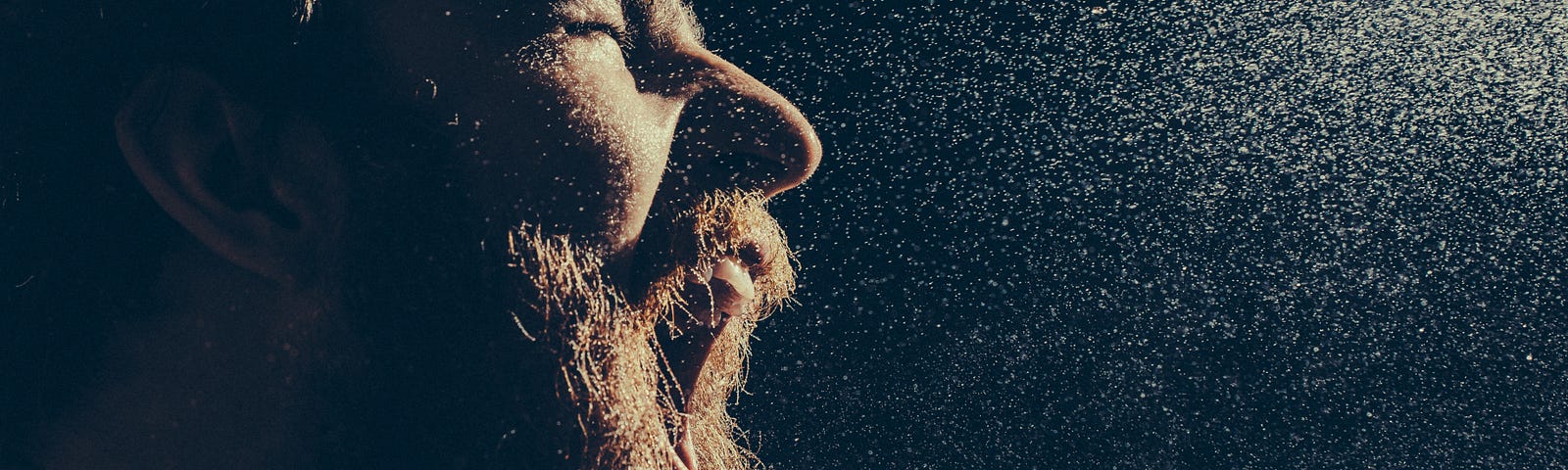 Naked bearded man angrily screams into a spray of water against a black background with copy space. Emotional portrait of man